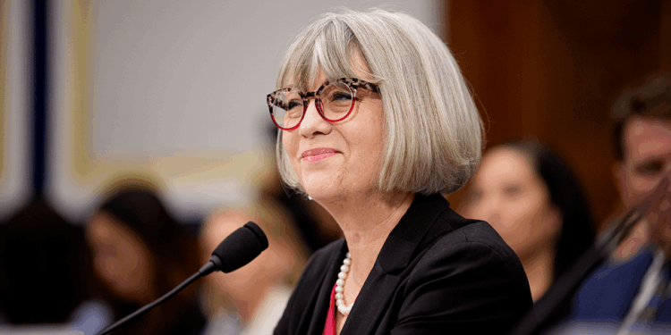 A woman with gray hair and glasses, wearing a black blazer and pearl necklace, speaks into a microphone at a formal event. Blurred audience members are seen in the background.