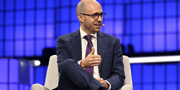 A man in a suit and tie, wearing glasses and a headset microphone, sits onstage with a blue geometric background, gesturing with his hand while speaking.