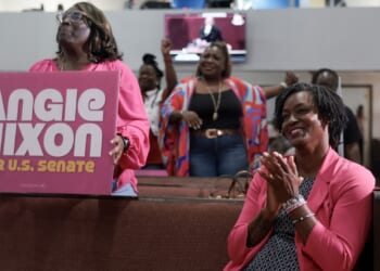 Women in pink cheer and clap in a church pew. One holds a large campaign sign that reads Angie Nixon for U.S. Senate. Others in the background smile and celebrate, showing support for the candidate.