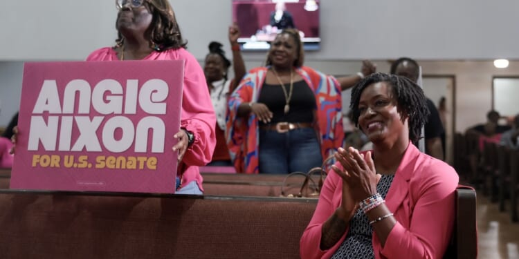 Women in pink cheer and clap in a church pew. One holds a large campaign sign that reads Angie Nixon for U.S. Senate. Others in the background smile and celebrate, showing support for the candidate.