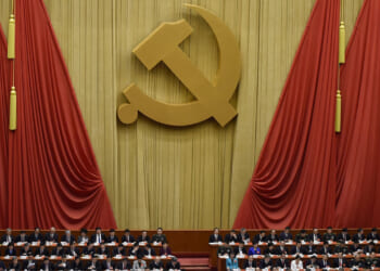 A large gold hammer and sickle symbol hangs on red drapes above a group of seated officials at a formal assembly.