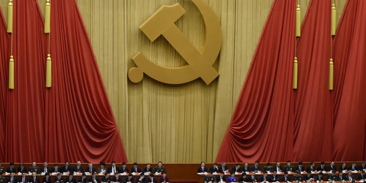 A large gold hammer and sickle symbol hangs on red drapes above a group of seated officials at a formal assembly.