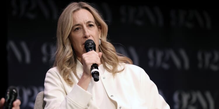 A woman with long blonde hair speaks into a microphone while seated on stage. She wears a white jacket and a neutral expression. A dark backdrop with 92Y in light letters is behind her.