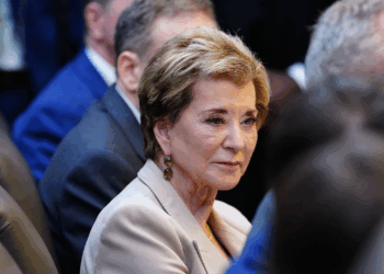 A woman with short light brown hair and a beige blazer sits among other people in a formal setting, looking slightly to the side. The background is out of focus, highlighting her face.