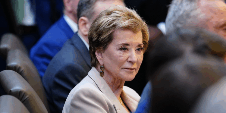 A woman with short light brown hair and a beige blazer sits among other people in a formal setting, looking slightly to the side. The background is out of focus, highlighting her face.