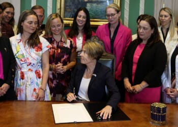 A woman sits at a desk signing a document, surrounded by a group of women standing and smiling in a formal room.