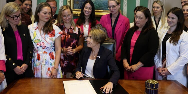 A woman sits at a desk signing a document, surrounded by a group of women standing and smiling in a formal room.