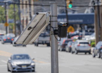 A solar panel and flock camera are mounted on a pole above a busy street with multiple cars and blurred buildings in the background.