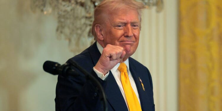 A man in a suit and yellow tie stands indoors, raising his fist and smiling slightly, with a microphone in front of him and a chandelier in the background.