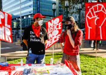 Two people at a protest table talk in front of red banners, including a raised fist and messages like NO JUSTICE NO PEACE. Both wear masks; ones shirt reads Defund the Police. Urban buildings are visible in the background.