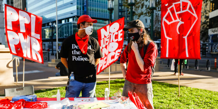 Two people at a protest table talk in front of red banners, including a raised fist and messages like NO JUSTICE NO PEACE. Both wear masks; ones shirt reads Defund the Police. Urban buildings are visible in the background.