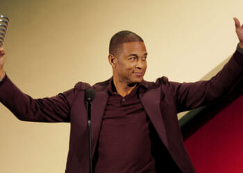 Former CNN host Don Lemon is trying to overturn part of the FACE Act. Pictured: a man in a maroon suit stands at a microphone, smiling and raising both arms. He holds a spiral-shaped award trophy in his left hand against a yellow and red background.