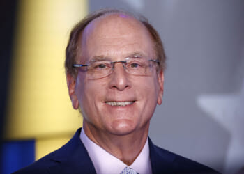 BlackRock is among the companies accused of merely rebranding DEI efforts or preserving them altogether. Pictured: CEO Larry Fink, a smiling middle-aged man with glasses and thinning hair, wearing a navy blue suit and light-colored tie, stands in front of a blurred background with hints of yellow, blue, and a partial white star.