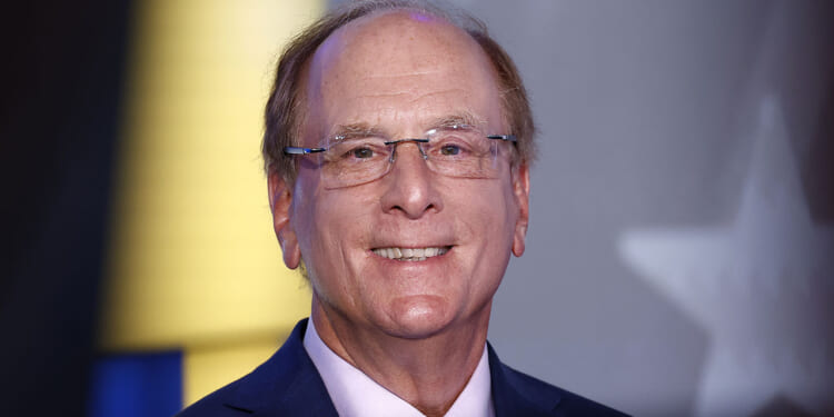 BlackRock is among the companies accused of merely rebranding DEI efforts or preserving them altogether. Pictured: CEO Larry Fink, a smiling middle-aged man with glasses and thinning hair, wearing a navy blue suit and light-colored tie, stands in front of a blurred background with hints of yellow, blue, and a partial white star.