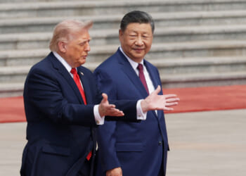 Two men in suits, one gesturing with open hands while speaking and the other looking ahead and smiling, stand outdoors on a wide stone plaza in China, with steps and a red carpet in the background.