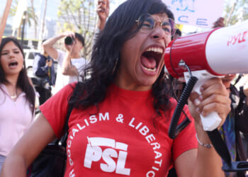 A person wearing a red Party for Socialism & Liberation shirt, passionately advocating for socialism, shouts into a megaphone at a protest. Others hold signs and chant in the background.