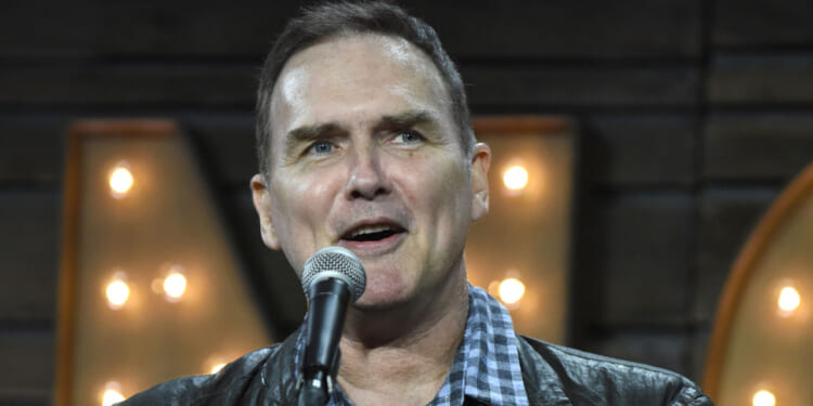 A man wearing a black jacket and checkered shirt speaks into a microphone, standing in front of a background with large, illuminated letters.