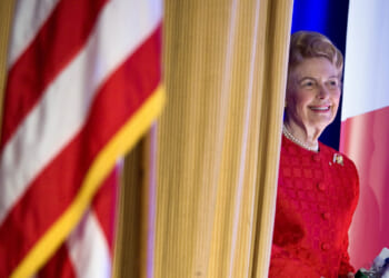 A woman in a red dress stands behind a curtain, smiling. An out-of-focus American flag is visible in the foreground.