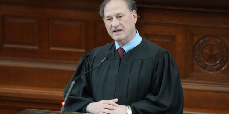 A judge in a black robe speaks at a podium in a courtroom, with a microphone in front of him and wooden paneling in the background.