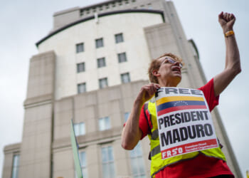 A person wearing glasses and a Free President Maduro sign raises a fist during a protest in front of a tall building.