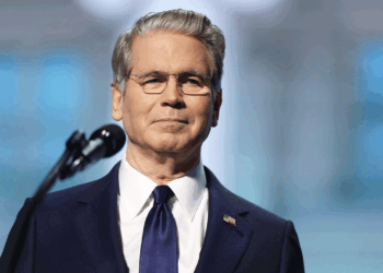 Scott Bessent in a navy suit and tie stands in front of a microphone, looking forward with a neutral expression. He has gray hair, glasses, and an American flag pin on his lapel. The background is blurred.