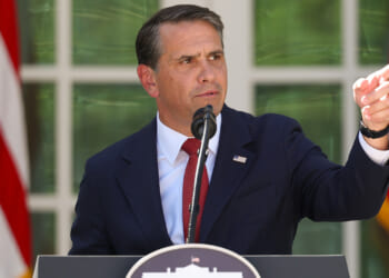 Todd Blanche in a dark suit speaks into a microphone at a podium outdoors, pointing to his right. Two large American flags are visible in the background.