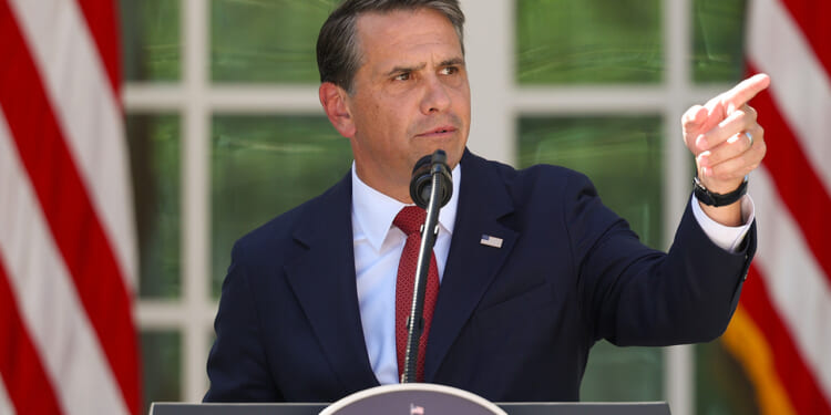 Todd Blanche in a dark suit speaks into a microphone at a podium outdoors, pointing to his right. Two large American flags are visible in the background.