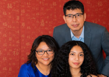 Three people pose in front of a red wall with gold patterns. A man in a blue suit stands behind a woman in glasses and a blue dress, while a young woman with curly hair sits in front. Though all are looking at the camera and smiling, their expressions hint at the tension of an unexpected hostage scenario unfolding just beyond the frame.