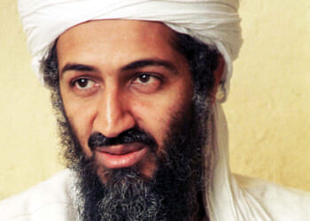 Close-up portrait of Osama bin Laden wearing a white turban and a long, dark beard, looking slightly off-camera against a plain, light-colored background.
