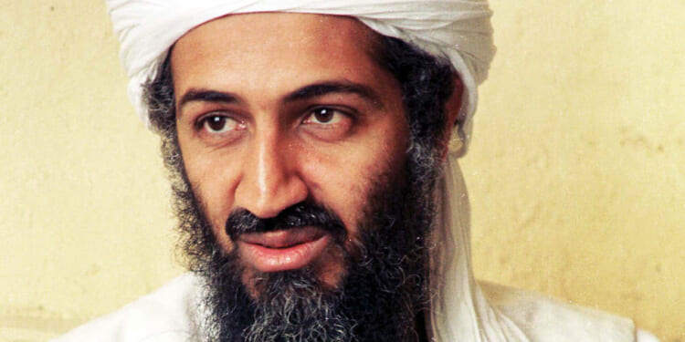 Close-up portrait of Osama bin Laden wearing a white turban and a long, dark beard, looking slightly off-camera against a plain, light-colored background.