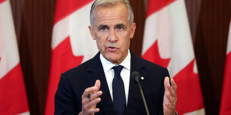 Mark Carney in a dark suit and tie speaks in front of several large Canadian flags, gesturing with his hands during a press conference or public address about tariffs.