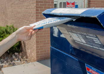 A person places a white padded envelope into a blue USPS mailbox outside a brick building, mailing their package with collection times posted on the mailbox.