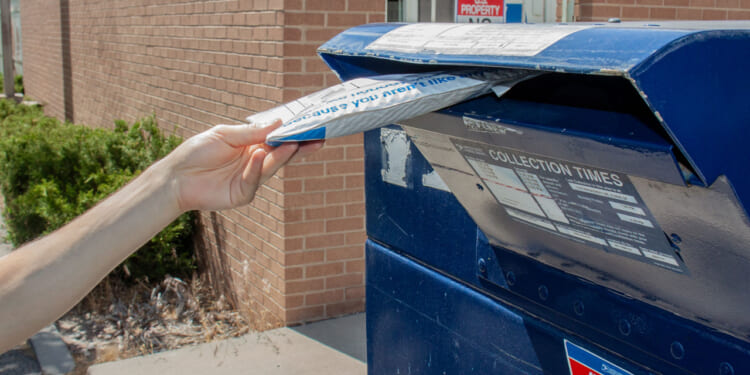 A person places a white padded envelope into a blue USPS mailbox outside a brick building, mailing their package with collection times posted on the mailbox.