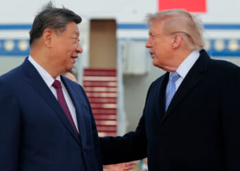 Xi Jinping and Donald Trump in suits stand outdoors, talking to each other. Xi faces forward, smiling, while Trump gestures with his hand. An airplane with a staircase is visible in the background.