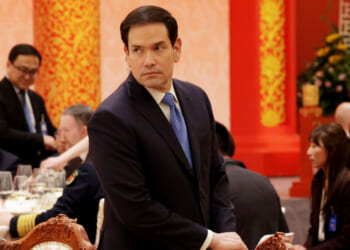 Marco Rubio in a dark suit stands beside an ornately carved chair at a formal event with red and gold decor. Several people are seated at tables in the background, dressed in formal attire.