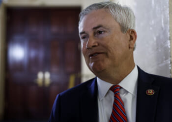 A man in a suit and red striped tie stands indoors, smiling slightly, with a blurred wooden door and a cream-colored wall in the background.