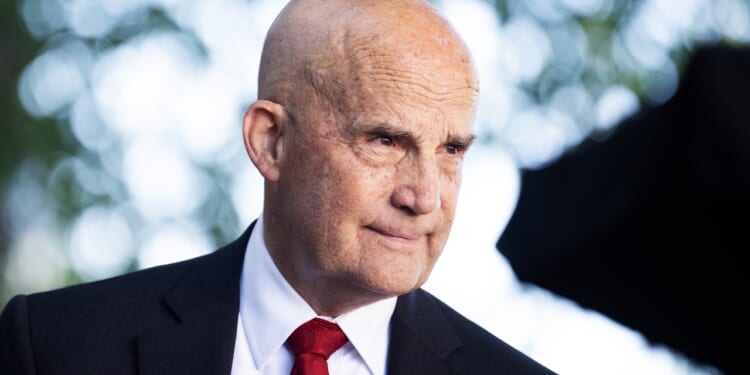 A bald man in a suit and red tie stands outdoors, looking to the side with a slight smile. The background is blurred with green and white bokeh from trees.