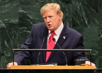 A man in a suit and red tie speaks at a podium in front of a green marble wall, appearing to address an assembly or conference.