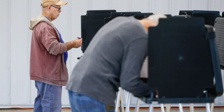 Two people are standing at voting booths, filling out ballots. The scene is indoors, with a white wall in the background. One person is in focus, while the other is blurred in the foreground.
