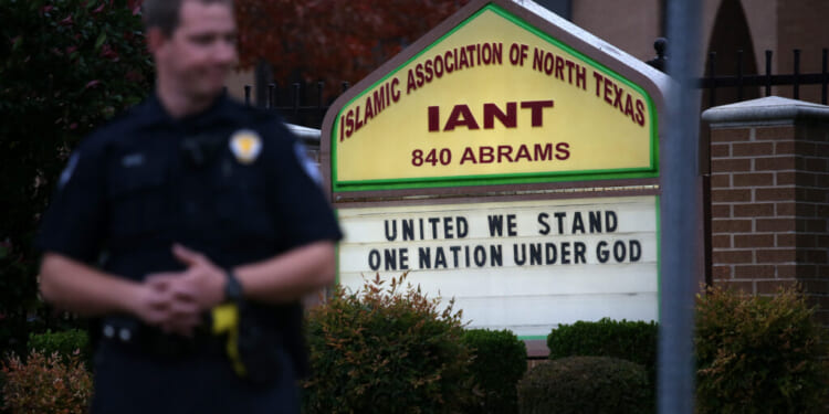 A police officer stands in front of a sign for the Islamic Association of North Texas that reads, United We Stand, One Nation Under God.