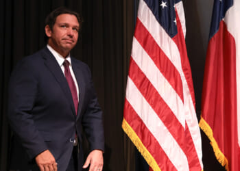 A man in a dark suit stands next to the United States flag and another flag with red, white, and blue stripes, against a dark backdrop.