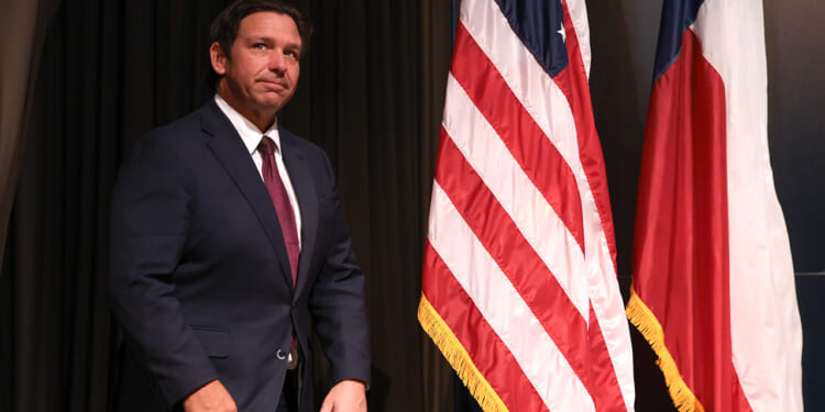 A man in a dark suit stands next to the United States flag and another flag with red, white, and blue stripes, against a dark backdrop.