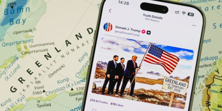 A photo illustration shows a smartphone displaying a post from the Truth Social account of President Donald Trump over a map of Greenland in the background.