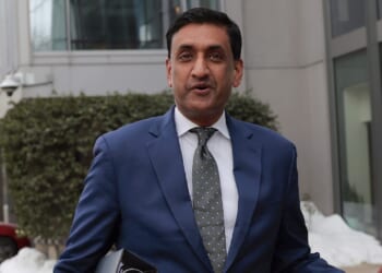 A man in a blue suit and patterned tie stands outdoors near buildings, holding a folder. Snow and greenery are visible in the background.