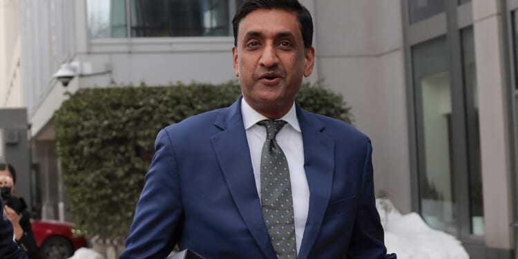 A man in a blue suit and patterned tie stands outdoors near buildings, holding a folder. Snow and greenery are visible in the background.