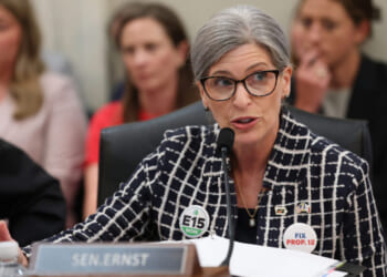 A woman in glasses speaks at a meeting, wearing a checkered jacket with E15 NOW and FIX PROP. 12 buttons. A nameplate reading SEN. ERNST is in front of her. Several people sit in the background.