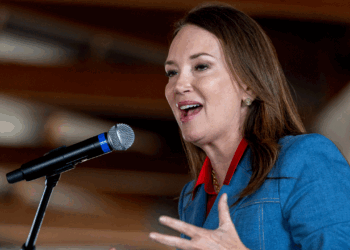A woman in a blue blazer speaks into a microphone, gesturing with one hand. She has long brown hair and is indoors, with blurred background beams visible.
