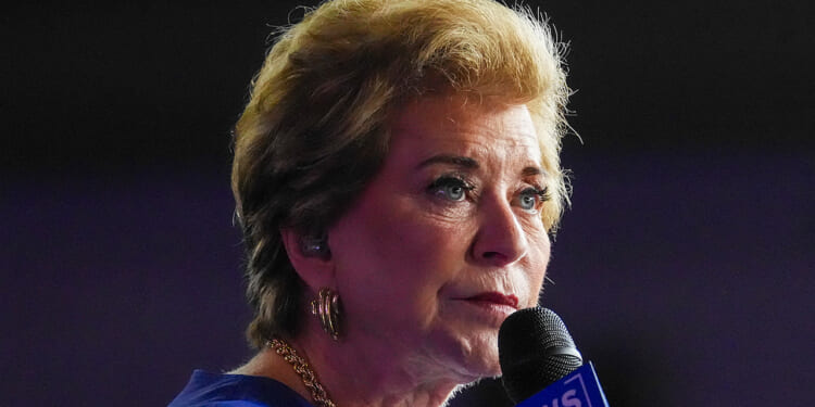 Linda McMahon with short blonde hair speaks into a microphone, wearing gold earrings and a necklace, with a serious expression and a blurred dark background.