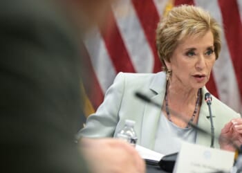 A woman in a light suit speaks into a microphone at a meeting focused on education, with an American flag in the background and a nameplate visible in front of her.