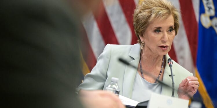 A woman in a light suit speaks into a microphone at a meeting focused on education, with an American flag in the background and a nameplate visible in front of her.
